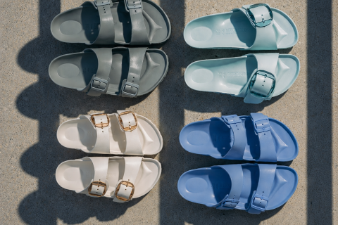 Vier Paar Sandalen in Grau, Hellblau, Creme und Blau sind auf Beton zu einem Quadrat angeordnet, über das Schatten fallen.