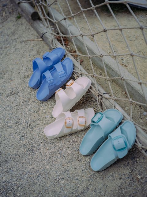 Drei Paar Sandalen mit Schnallen – blau, weiß und hellblau – liegen auf sandigem Boden und lehnen an einem Netz.