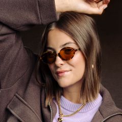 Eine Frau mit braunen Haaren, die eine Sonnenbrille mit Schildpatt-Rahmen, eine braune Jacke, ein lila Oberteil und goldene Halsketten trägt und lächelnd einen Arm hebt.