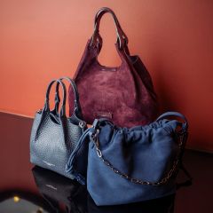 Drei stilvolle Handtaschen – eine in Burgunderrot, eine in Marineblau mit einer Kette und eine in Dunkelgrau – wurden vor einer rötlichen Wand auf einer spiegelnden Oberfläche präsentiert.