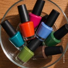 Eine Glasschale, gefüllt mit sieben Nagellackfläschchen in verschiedenen Farben, darunter Blau, Rosa, Orange, Grün, Blaugrün, Limettengrün und Braun.