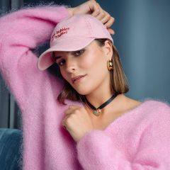 Eine Frau in einem flauschigen rosa Pullover und einer rosa Mütze posiert in einem Raum; sie trägt goldene Ohrringe und ein schwarzes Halsband mit einem türkisfarbenen Herzanhänger.