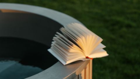 Ein aufgeschlagenes Buch, dessen Seiten aufgefächert sind, liegt am Rand einer runden Wanne im Freien, im Hintergrund ist grünes Gras zu sehen.