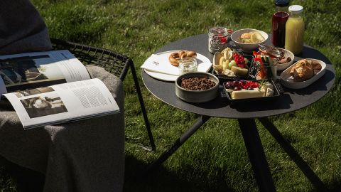Ein runder Tisch auf dem Rasen mit verschiedenen Frühstücksspeisen, Getränken und einer aufgeschlagenen Zeitschrift auf einem Stuhl in der Nähe, der mit einer Decke bedeckt ist.
