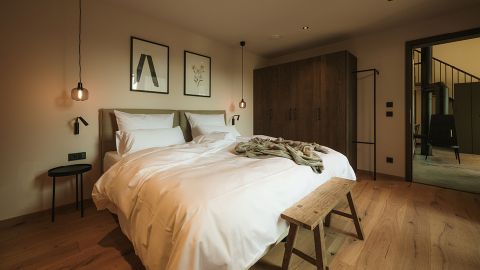 Ein gemütliches Schlafzimmer mit einem Doppelbett, weißer Bettwäsche, einer Holzbank, einem Kleiderschrank, gerahmten Bildern und warmer Beleuchtung vor hellen Wänden und einem Holzboden.