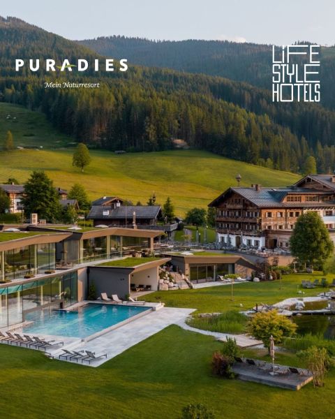 Modernes Resort mit Außenpool in einem grünen Tal, umgeben von traditionellen Holzgebäuden und bewaldeten Hügeln unter strahlend blauem Himmel.