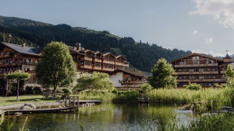 Holzhütten stehen an einem kleinen Teich, der von Grün umgeben ist, mit bewaldeten Hügeln und einem blauen Himmel im Hintergrund.