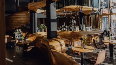 Ein modernes Café mit einer geschwungenen Holztheke, runden Tischen, hellbraunen Stühlen, Hängelampen und großen Fenstern, durch die natürliches Sonnenlicht hereinströmt.