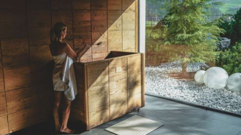 Im Innenraum steht eine Frau in einem Handtuch neben einer Holzbadewanne, während Sonnenlicht durch große Fenster mit Blick auf einen Garten hereinströmt.