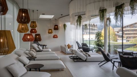 Moderne Lounge mit weißen Liegesesseln, geometrischen Pendelleuchten, Pflanzen und großen Fenstern mit Blick auf einen Außenbereich mit Gebäuden und Bäumen.