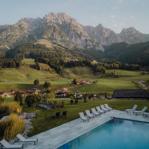 Ein Swimmingpool mit Liegestühlen bietet einen Blick auf ein grasbewachsenes Tal, vereinzelte Häuser und hohe, grüne Berge unter einem klaren Himmel.