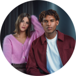 Zwei Personen posieren in einem Raum, eine trägt einen rosa Pullover, die andere eine weinrote Jacke; beide blicken direkt in die Kamera.