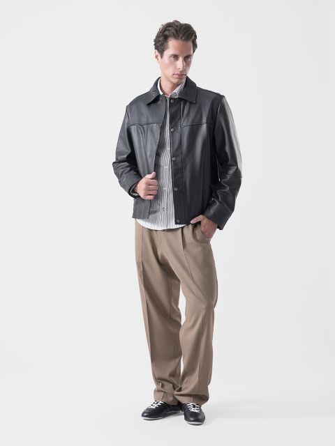 Ein Mann, der eine schwarze Lederjacke, ein gestreiftes Hemd, eine hellbraune Hose und schwarze Turnschuhe trägt, steht vor einem schlichten weißen Hintergrund und blickt leicht zur Seite.