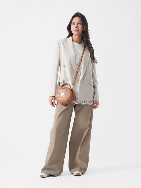 Eine Frau, die einen beigen Blazer, ein weißes Oberteil, eine hellbraune Hose mit weitem Bein, Sandalen und eine runde, hellbraune Umhängetasche trägt, steht vor einem schlichten weißen Hintergrund.