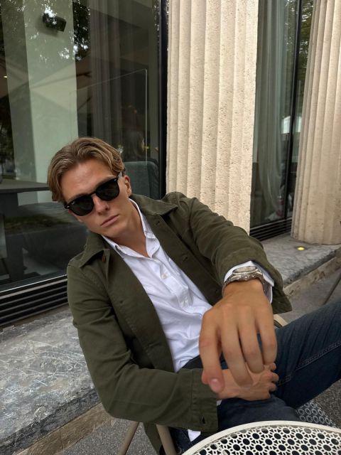 Ein Mann mit Sonnenbrille und grüner Jacke sitzt an einem Tisch im Freien, lehnt sich zurück und streckt einen Arm in Richtung Kamera aus.