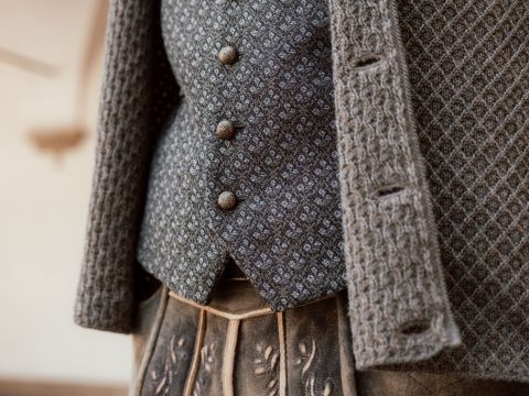 Nahaufnahme einer Person, die eine gemusterte Weste, eine strukturierte Strickjacke und einen bestickten Rock oder eine bestickte Hose in Braun- und Grautönen trägt.