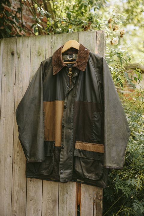 Eine Patchwork-Jacke mit einem Kragen aus Cord hängt draußen an einem Holzzaun, umgeben von Grünpflanzen und Blumen.