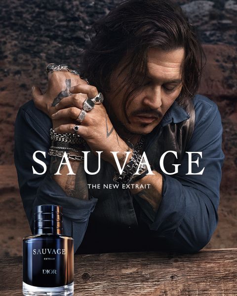 Ein Mann mit Tattoos und Schmuck lehnt sich an einem Tisch neben einer Flasche „Dior Sauvage Extrait“-Parfüm vor einer rauen Außenkulisse.