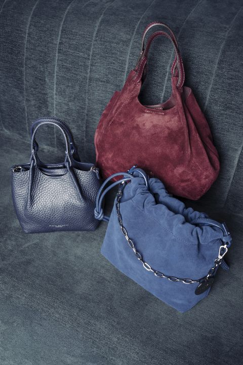 Three handbags on dark blue denim: a burgundy suede tote, a blue pebbled purse, and a blue bucket bag with a chain strap.