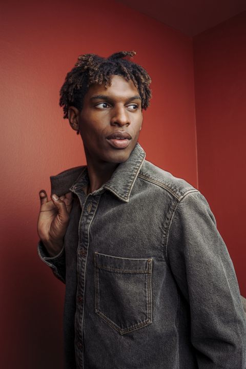Young man in a denim jacket looking to the right against a red wall.
