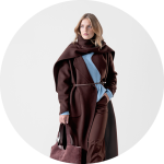 Eine Frau in einem weinroten Mantel mit einem blauen Schal und einer weinroten Hose, die eine dazu passende Tasche in der Hand hält und vor einem schlichten weißen Hintergrund posiert.