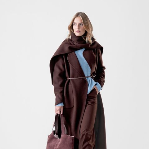 Eine Frau in einem langen braunen Mantel und einem blauen Pullover, die eine große braune Tasche in der Hand hält und vor einem schlichten weißen Hintergrund steht.