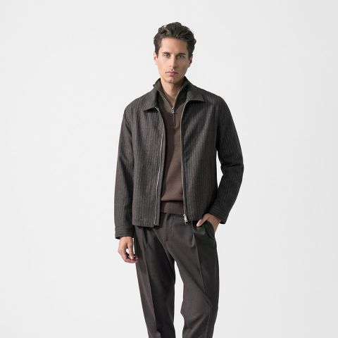 Ein Mann, der eine braune Jacke mit Reißverschluss über einem braunen Hemd und einer dazu passenden braunen Hose trägt und vor einem schlichten weißen Hintergrund steht.
