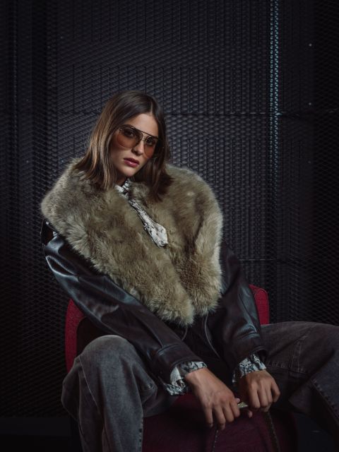 Eine Frau mit Sonnenbrille und Lederjacke mit großem Pelzkragen sitzt auf einem roten Stuhl vor einem dunklen, strukturierten Hintergrund und blickt selbstbewusst in die Kamera.