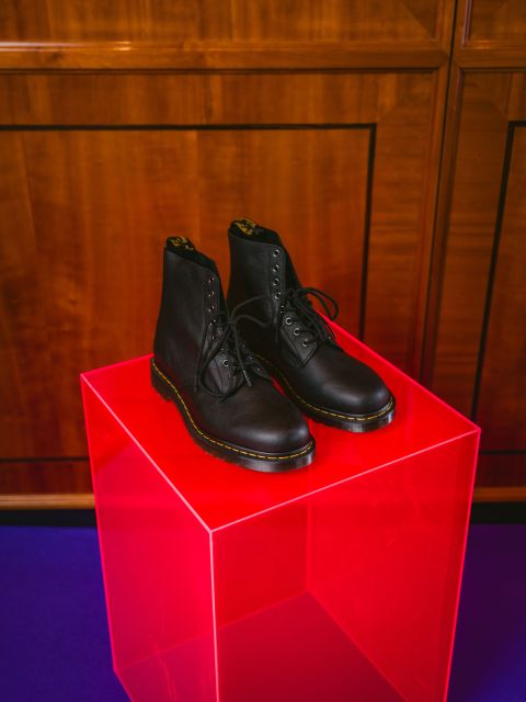 Schwarze Schnürstiefel auf einem leuchtend pinkfarbenen, durchsichtigen Würfel, vor einem Hintergrund aus Holzpaneelen und auf einem blauen Teppichboden.