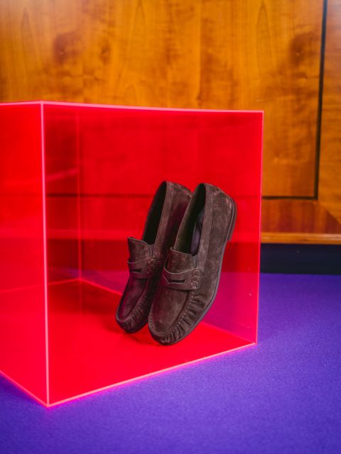 Ein Paar dunkle Wildleder-Loafer, präsentiert in einer durchsichtigen roten Schachtel auf einer violetten Unterlage, mit einer Holzwand im Hintergrund.