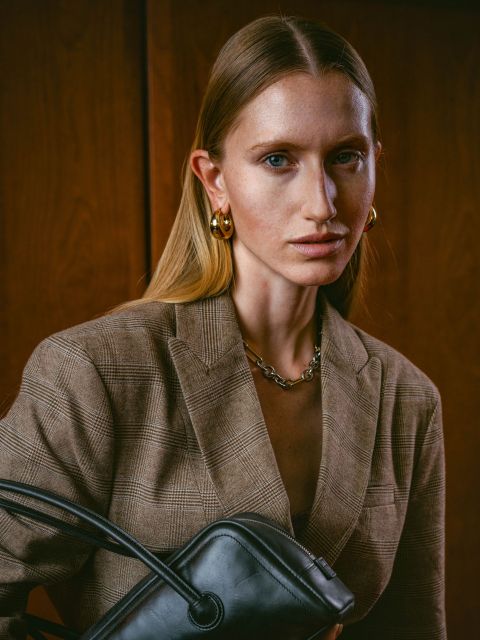 Eine Frau in einem braunen karierten Blazer hält eine schwarze Handtasche in der Hand, steht vor Holzvertäfelungen und trägt goldene Ohrringe sowie eine Kettenhalskette.
