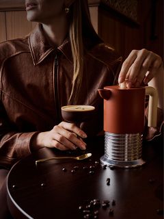 Eine Frau in einer braunen Jacke sitzt an einem Tisch, auf dem ein Kaffee-Cocktail, eine French Press und verstreute Kaffeebohnen stehen.