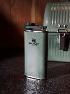 Auf einer dunklen Arbeitsplatte steht eine grüne Stanley-Thermosflasche, im Hintergrund ist vor einer Holzwand eine dazu passende grüne Brotdose zu sehen.