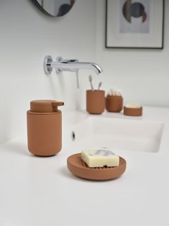 Ein modernes Waschbecken mit braunem Seifenspender, Seifenschale und Accessoires auf einer weißen Arbeitsplatte sowie einer darüber angebrachten Wandarmatur.