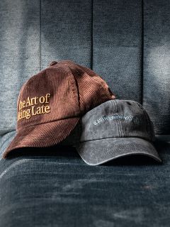 Zwei Mützen auf einem blauen Stoffsessel; eine braune mit der Aufschrift „The Art of Being Late“, die andere in verblasstem Schwarz mit kleinerer, schwerer lesbarer Schrift.