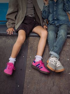 Zwei Kinder sitzen nebeneinander auf einer Rampe; man sieht nur ihre Beine und Turnschuhe – das eine trägt rosa Schuhe, das andere graue Turnschuhe und Jeans.