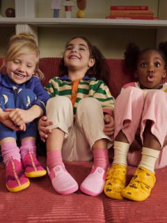 Drei kleine Kinder sitzen auf einem roten Sofa, lächeln und lachen; jedes trägt bunte Kleidung und unterschiedliche, farbenfrohe Schuhe.