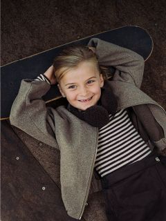 Ein lächelndes Kind in einem gestreiften Hemd und einer Jacke liegt auf einem Skateboard, die Hände hinter dem Kopf verschränkt, und blickt in die Kamera.