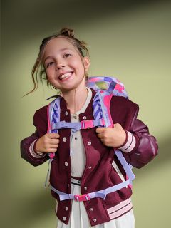 Ein lächelndes Mädchen mit einem Rucksack und einer weinroten Jacke steht vor einem grünen Hintergrund und sieht fröhlich und bereit für die Schule aus.