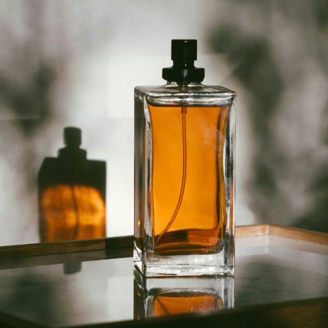 Ein Parfümflakon aus Glas mit bernsteinfarbener Flüssigkeit steht auf einem verspiegelten Tablett und wirft einen Schatten auf eine strukturierte, graue Wand.