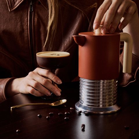 Eine Person in einer braunen Jacke hält ein Kaffeegetränk in der Hand und benutzt eine moderne, rote Kaffeekanne auf einem dunklen Tisch, auf dem ein goldener Löffel und verstreute Kaffeebohnen liegen.
