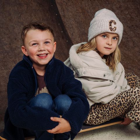 Zwei Kinder sitzen nebeneinander; das eine lächelt in einer blauen Jacke, das andere trägt einen weißen Hut und einen Mantel sowie eine Hose mit Leopardenmuster, beide posieren vor einem braunen Hintergrund.