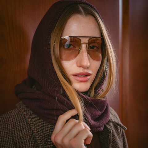 Eine Frau mit einer großen Sonnenbrille, einer burgunderroten Schal-Kapuze und einem braunen Karomantel steht vor einem hölzernen Hintergrund.