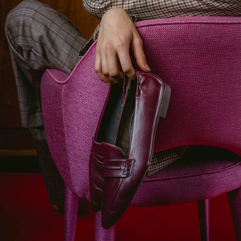 Eine Person in karierten Hosen sitzt auf einem rosa Stuhl und hält einen burgunderroten Slipper über die Rückenlehne des Stuhls.