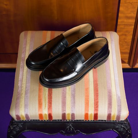 Ein Paar glänzende schwarze Slipper stehen auf einem gestreiften, gepolsterten Hocker in Orange-, Lila- und Beigetönen.