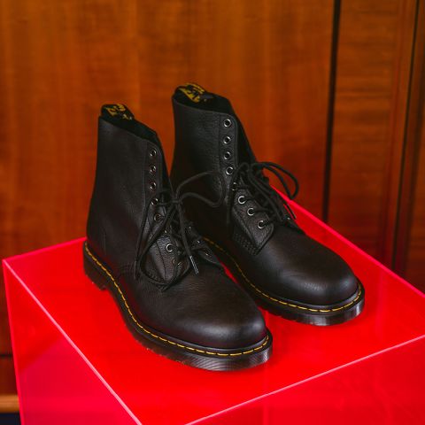 Ein Paar schwarze Lederstiefel mit Schnürung und gelben Ziernähten steht auf einem glänzenden roten Ausstellungsständer vor einem hölzernen Hintergrund.