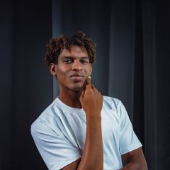 Ein Mann in einem weißen T-Shirt steht vor einem dunklen Hintergrund und wirkt nachdenklich, während er eine Hand an sein Kinn legt.
