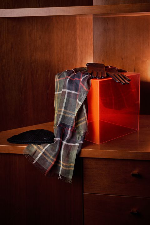 Ein karierter Schal und braune Handschuhe liegen über einer roten, durchscheinenden Schachtel auf einem Holzschrank, daneben steht ein schwarzer Hut.