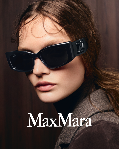 Eine Frau mit einer großen schwarzen Sonnenbrille und einem braunen Mantel vor einem dunklen Holzhintergrund, unten ist das „MaxMara“-Logo zu sehen.