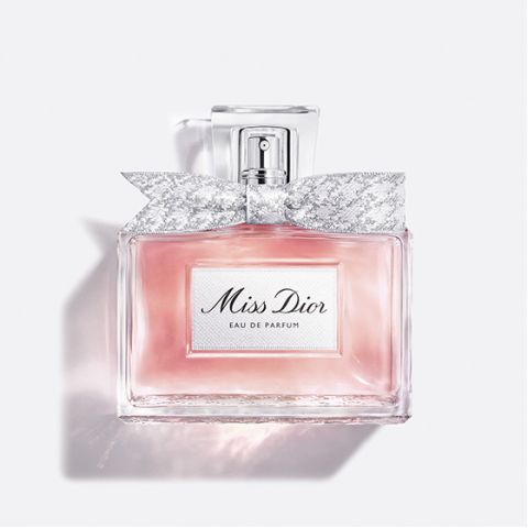 Eine durchsichtige Glasflasche mit „Miss Dior Eau de Parfum“, gefüllt mit rosa Flüssigkeit und einer silbernem, gemusterten Schleife um den Flaschenhals, vor einem schlichten weißen Hintergrund.